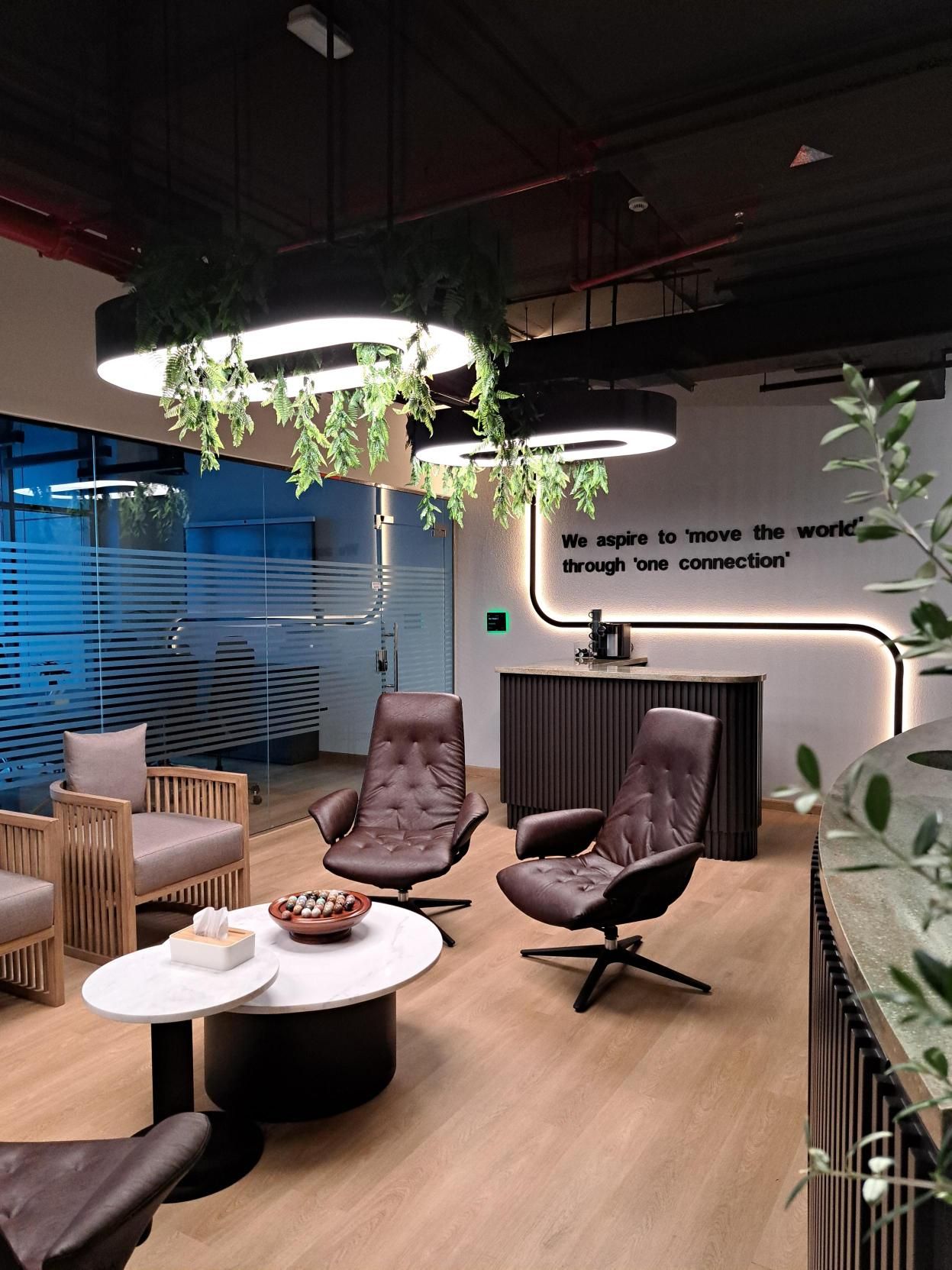 Office lounge with contemporary seating, round coffee tables, hanging greenery lights, and a wall quote.