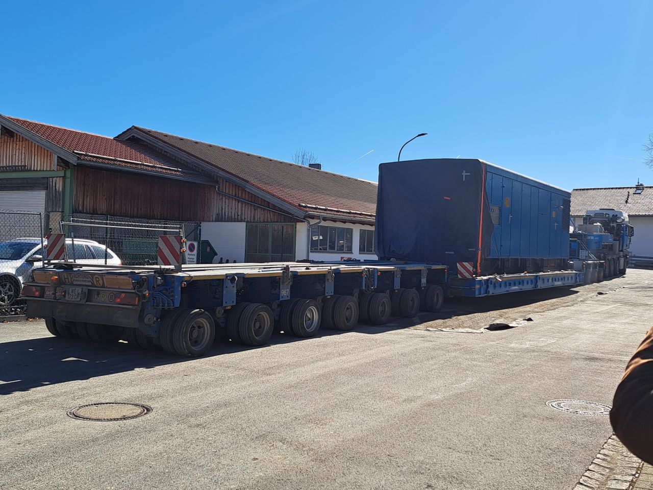 Large blue generator wrapped in protective cover, secured on a heavy-duty modular trailer being hauled by a truck through Hausham.