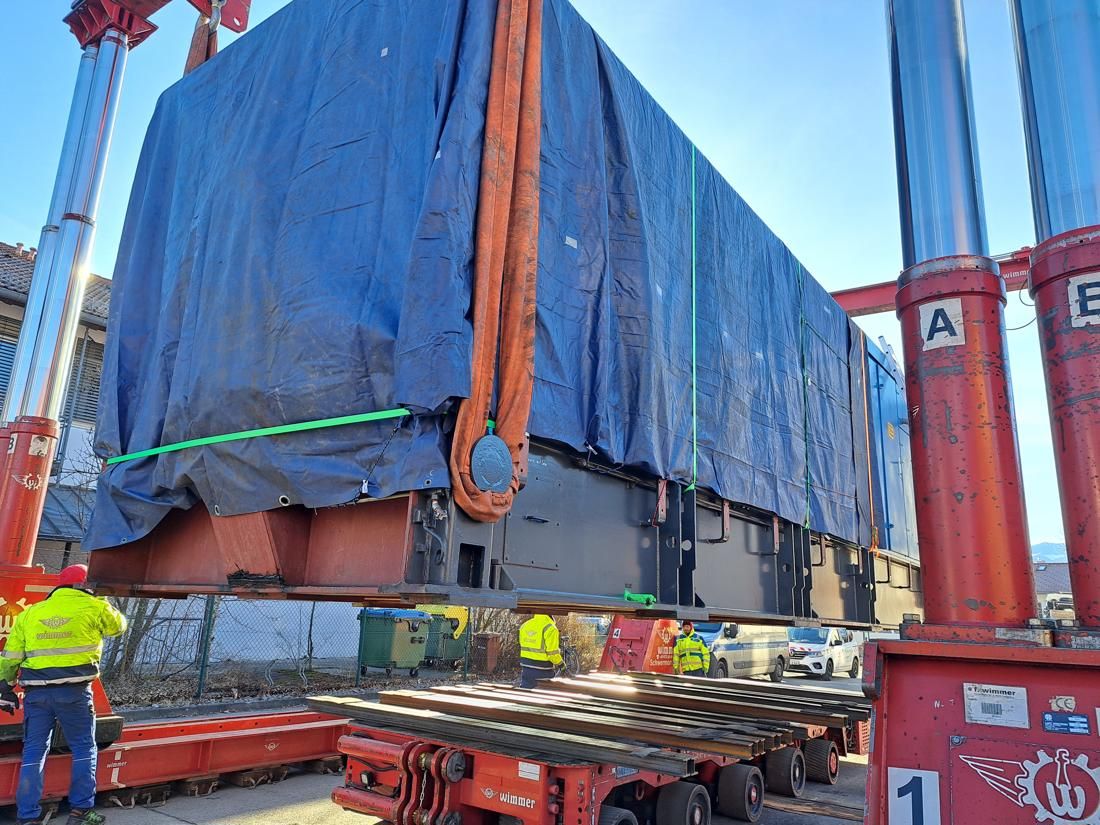 Generator wrapped in a blue tarp being lifted by a red gantry crane onto a multi-axle trailer.