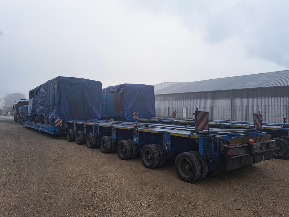 Two blue generator units covered with tarps on extended multi-axle heavy transport trailers.