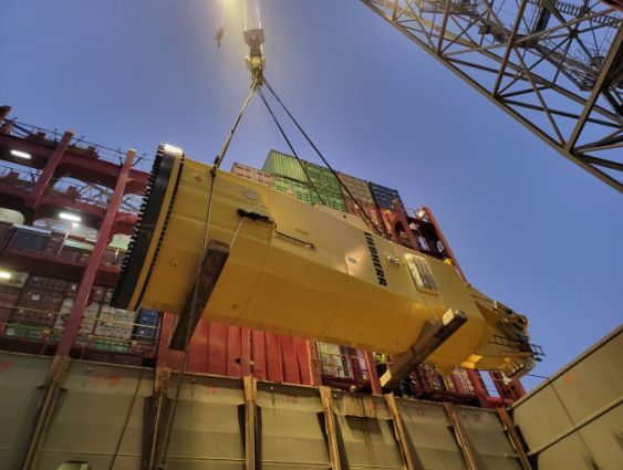 Yellow Liebherr slewing column being lifted by crane into a cargo ship’s hold at night, with container stacks in the background.