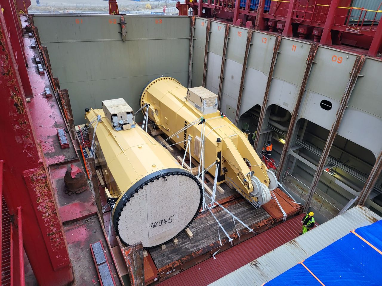Two large yellow Liebherr slewing columns lashed and stowed inside a cargo ship’s hold, viewed from above.
